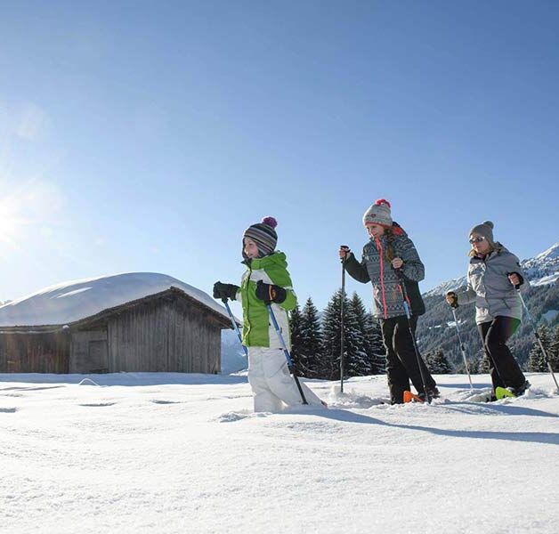 Eine Mutter geht mit ihrer Tochter und ihrem Sohn Schneeschuhwandern. Sie gehen im Tiefschnee an einer Hütte vorbei.