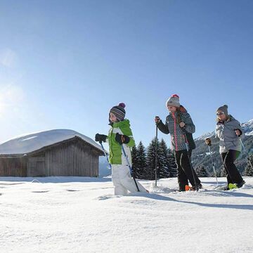 Eine Mutter geht mit ihrer Tochter und ihrem Sohn Schneeschuhwandern. Sie gehen im Tiefschnee an einer Hütte vorbei.