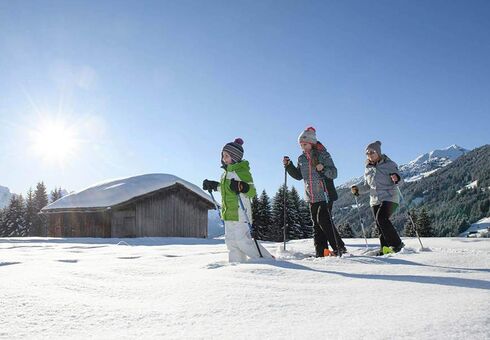 Eine Mutter geht mit ihrer Tochter und ihrem Sohn Schneeschuhwandern. Sie gehen im Tiefschnee an einer Hütte vorbei.