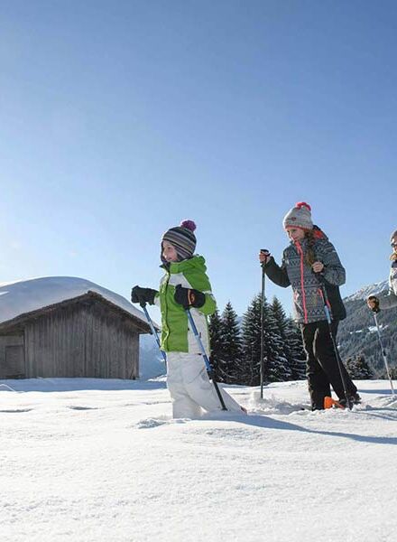 Eine Mutter geht mit ihrer Tochter und ihrem Sohn Schneeschuhwandern. Sie gehen im Tiefschnee an einer Hütte vorbei.